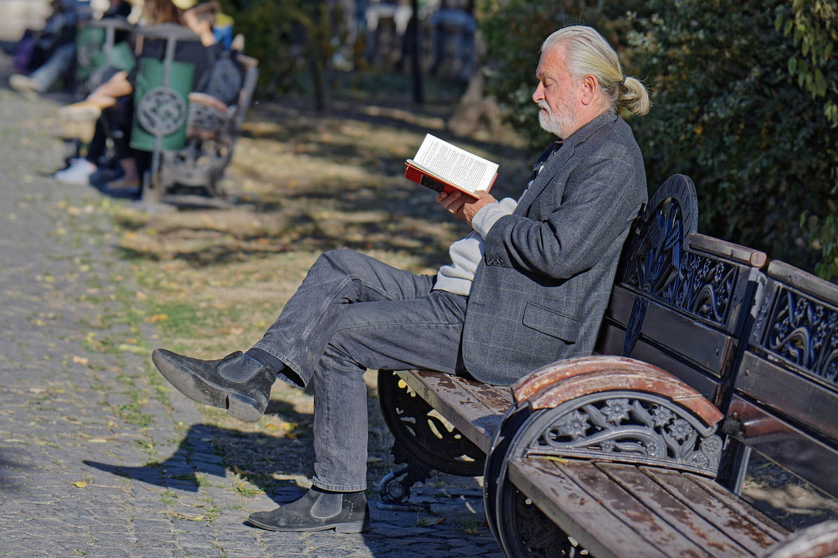 Äldre man sitter på en parkbänk i solen och läser en bok
